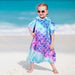 Child wearing mermaid scale hooded poncho towel in turquoise purple gradient on sandy beach